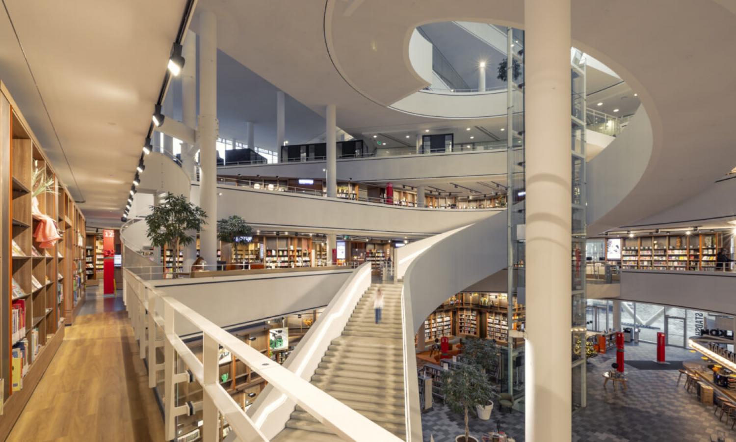 Bibliotheek Nieuwegein interieur. Trappenhuis met lift. Rekken met boeken. Design witte balustrades. Warm licht.
